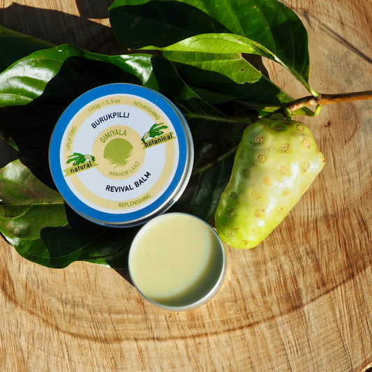 A tin of Burukpili Revival Salve shown open next to a small dish of pale yellow balm, styled with green leaves and a greenish-yellow fruit on a rustic wooden slice background.
