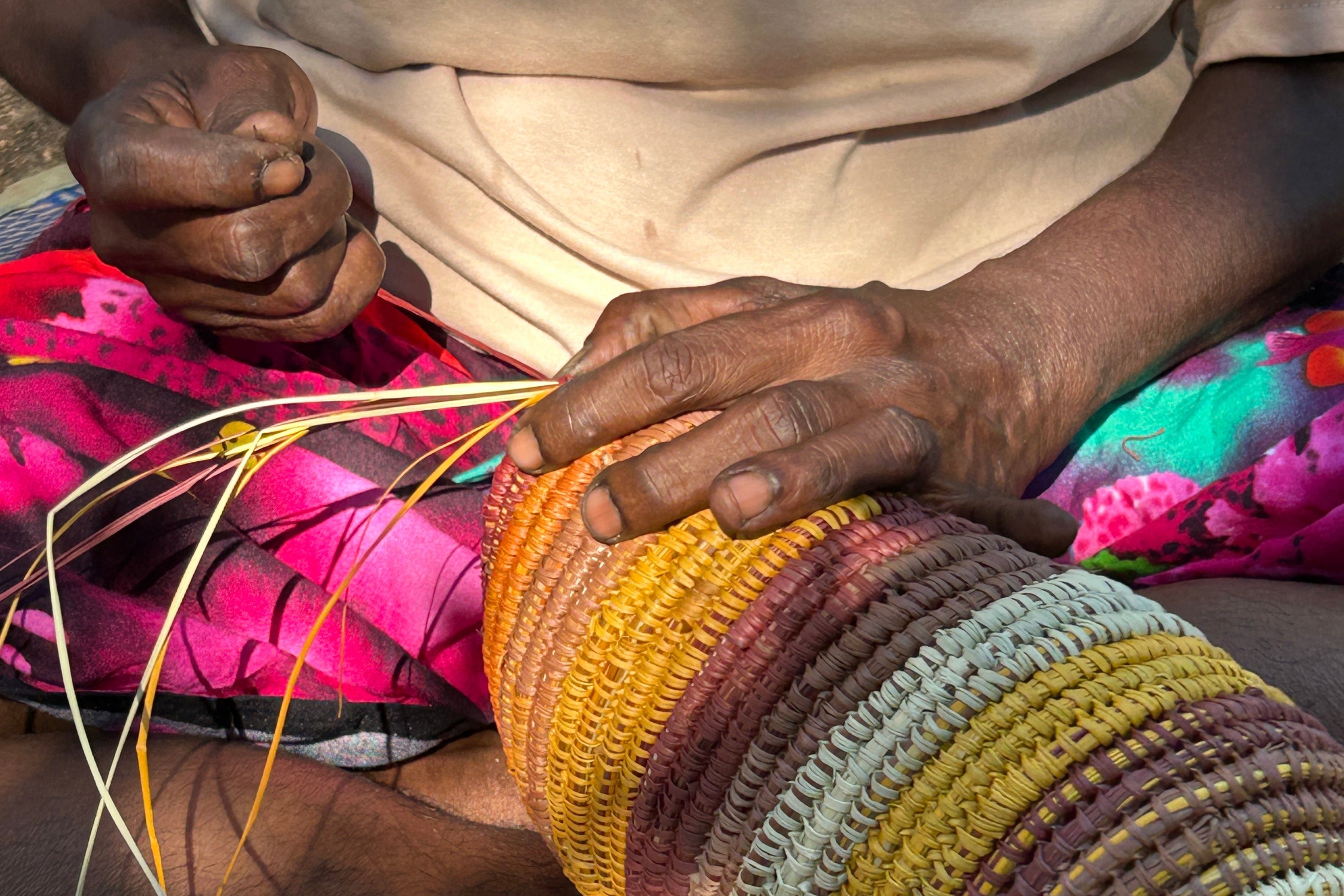 A close-up of a person's hands weaving a large, cylindrical basket or roll of woven material with multiple horizontal stripes in yellow, orange, brown, green, and blue. Thin string threads are being manipulated, and a pair of scissors lies on the patterned mat below.